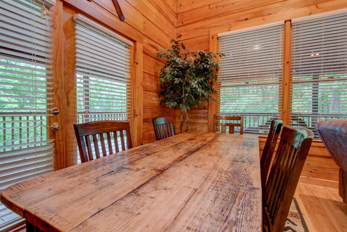 A large wooden dining table surrounded by chairs, with a lush green plant as a centerpiece. Sliding glass doors lead to a deck, suggesting a seamless indoor-outdoor flow. The setting is warm and intimate, perfect for group meals, celebrations, and conversations.