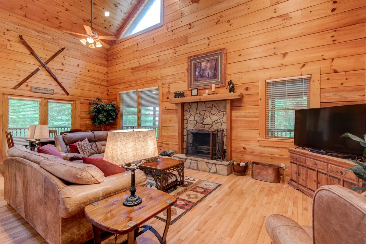A spacious, open living area featuring a grand stone fireplace, large windows with views of the surrounding greenery, exposed wooden beams, and a comfortable seating arrangement. The room is designed for gatherings, with ample space for family games, movie nights, or simply relaxing by the fire.
