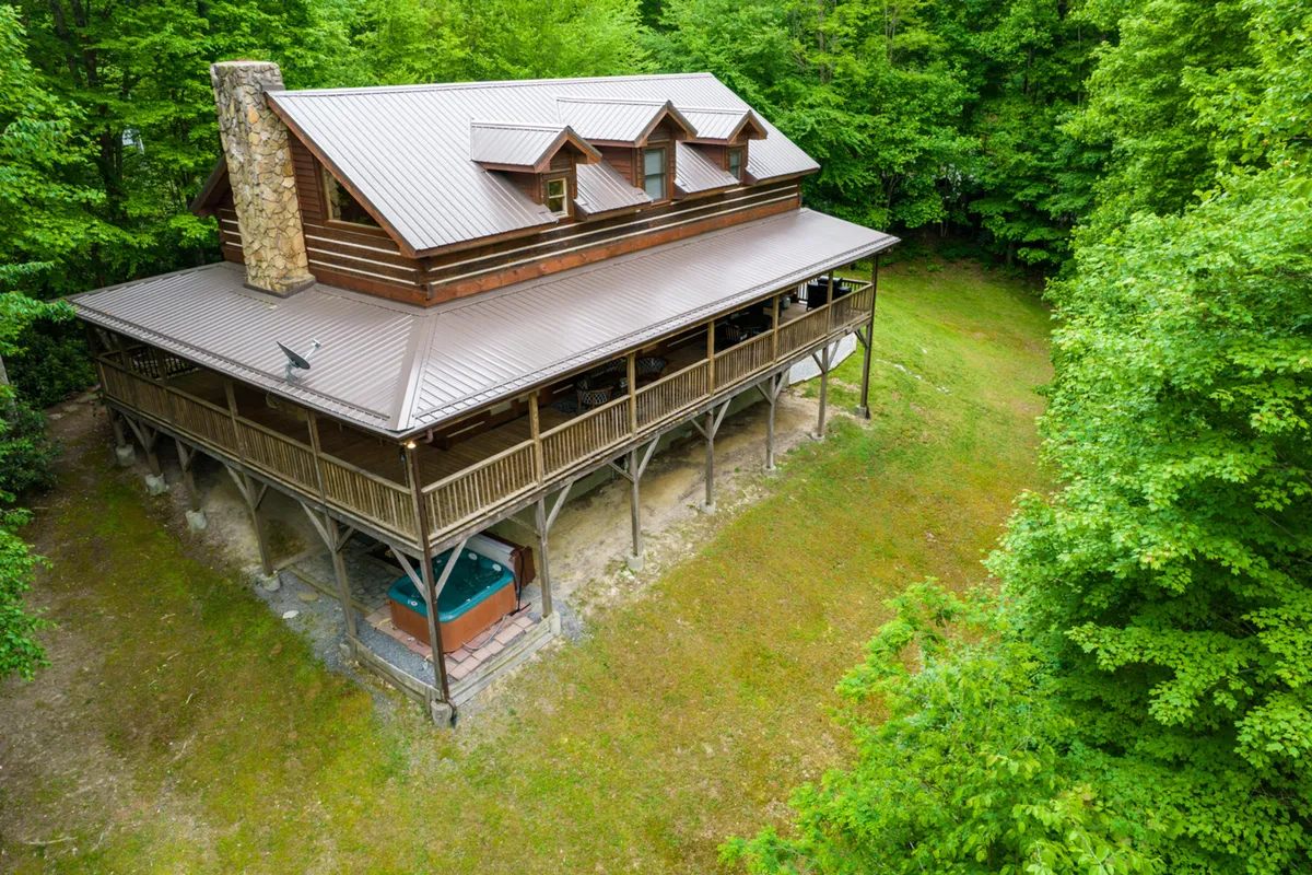 This exterior shot captures the large wooden cabin surrounded by tall trees, featuring a spacious wraparound porch with seating and an inviting atmosphere. The porch area includes an outdoor hot tub and hints of spaces ideal for group gatherings, barbecues, and relaxation amidst nature.
