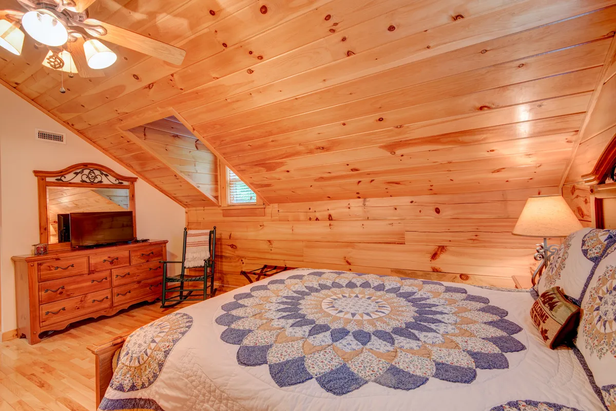 A cozy bedroom with a quilted bedspread, wooden dresser, ceiling fan, and a charming rocking chair. Soft, welcoming lighting and rustic decor create an inviting ambiance ideal for relaxation.