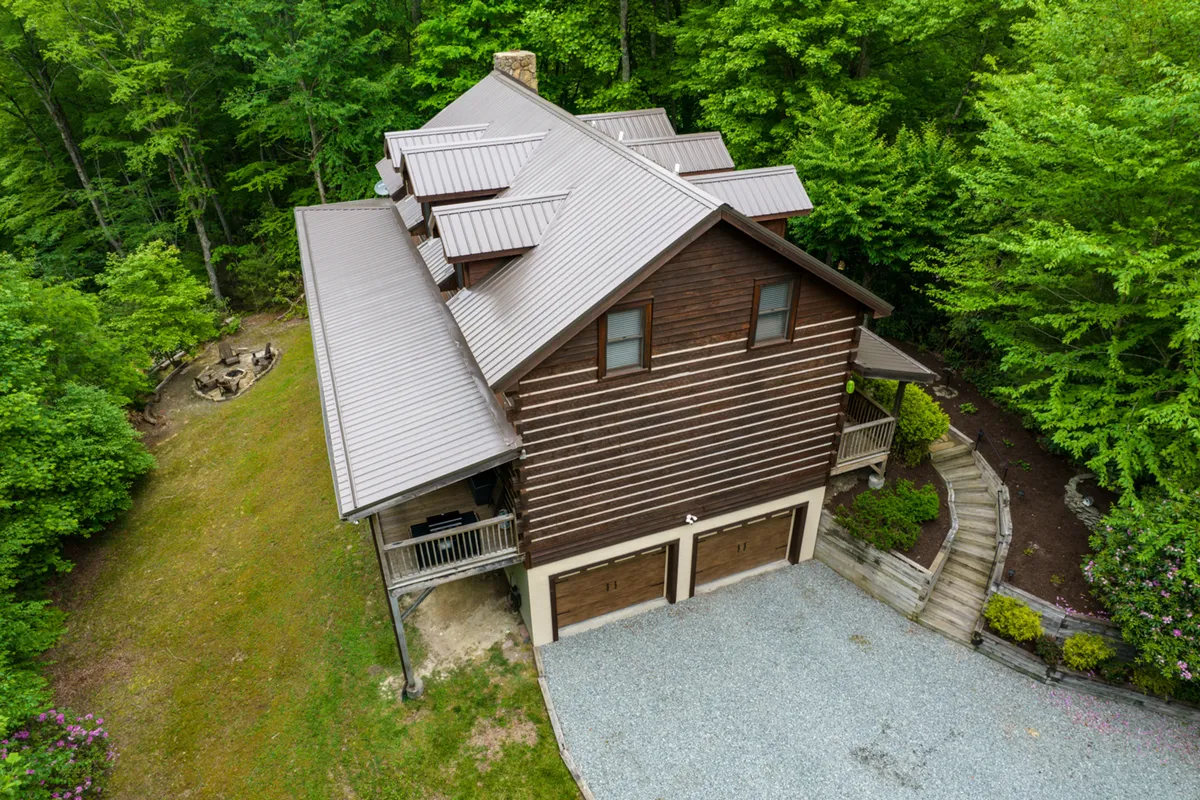 A welcoming exterior shot featuring the wooden house nestled among lush greenery, with a sloped yard and a dedicated fire pit area visible. The open lawn in the foreground provides ample space for pets and families to play and explore.