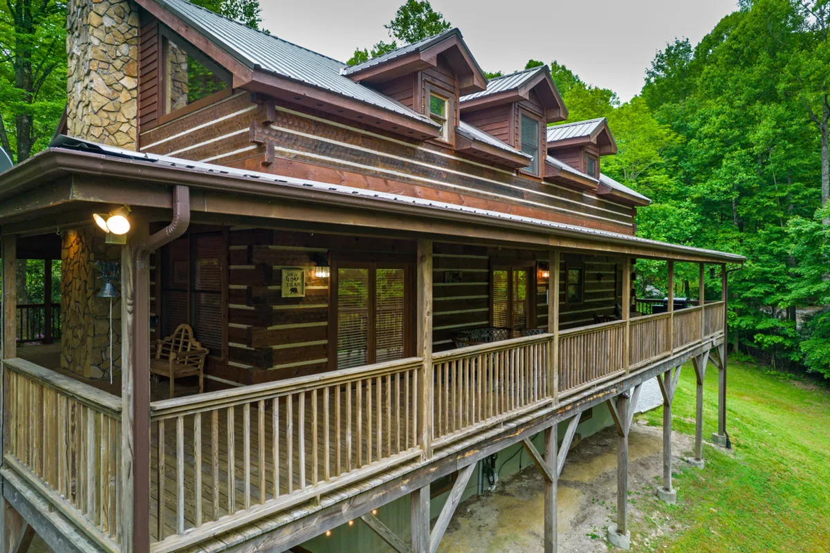 This wide-angle exterior image presents the cabin’s log structure, welcoming wooden porch, and abundant greenery. The image gives a sense of place and setting—showing the home’s private yet accessible location near both nature and local attractions. The porch is visible as a gathering place, supporting the idea of outdoor meals, coffee, and relaxation.