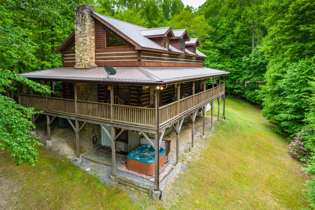 The image showcases a classic log cabin nestled among tall, lush trees, with a spacious wraparound porch and a bubbling hot tub on the deck. The setting feels private and serene, with inviting outdoor spaces perfect for relaxation or social gatherings.