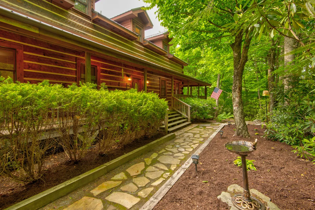 A charming log cabin exterior with a stone pathway, surrounded by greenery, an American flag, and a birdbath. The inviting porch and garden areas offer multiple points for exploration, relaxation, and play.