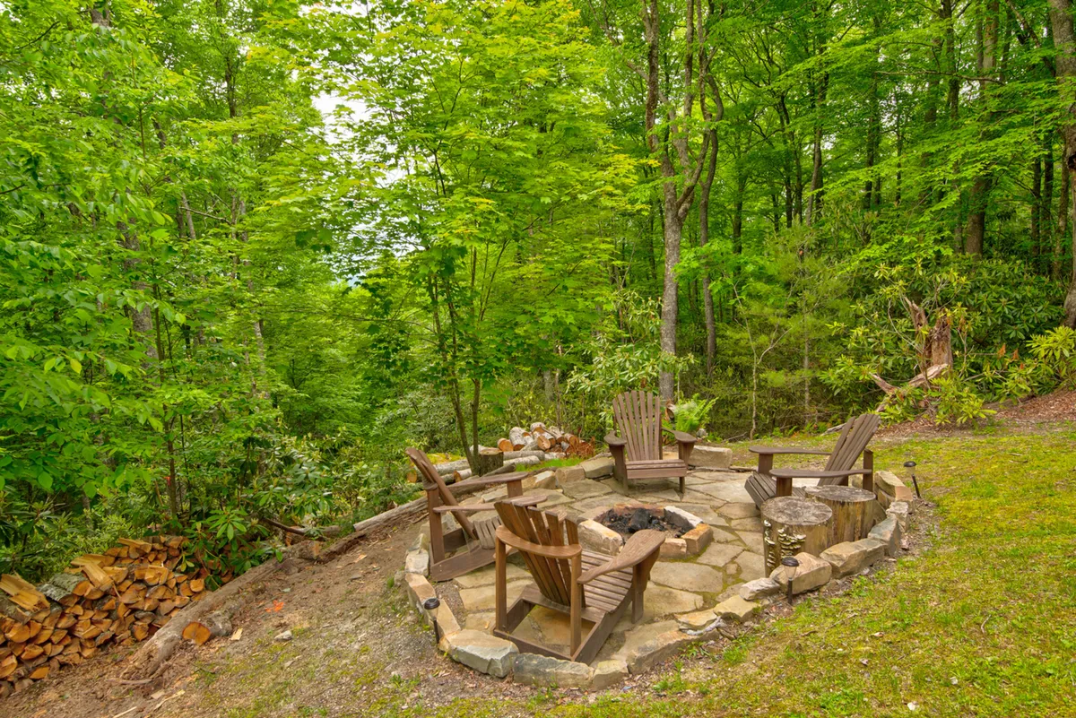 A cozy outdoor fire pit area with wooden chairs arranged for gatherings, set amidst a backdrop of natural greenery. The setup is casual and welcoming, ideal for evenings spent outdoors with pets and family.