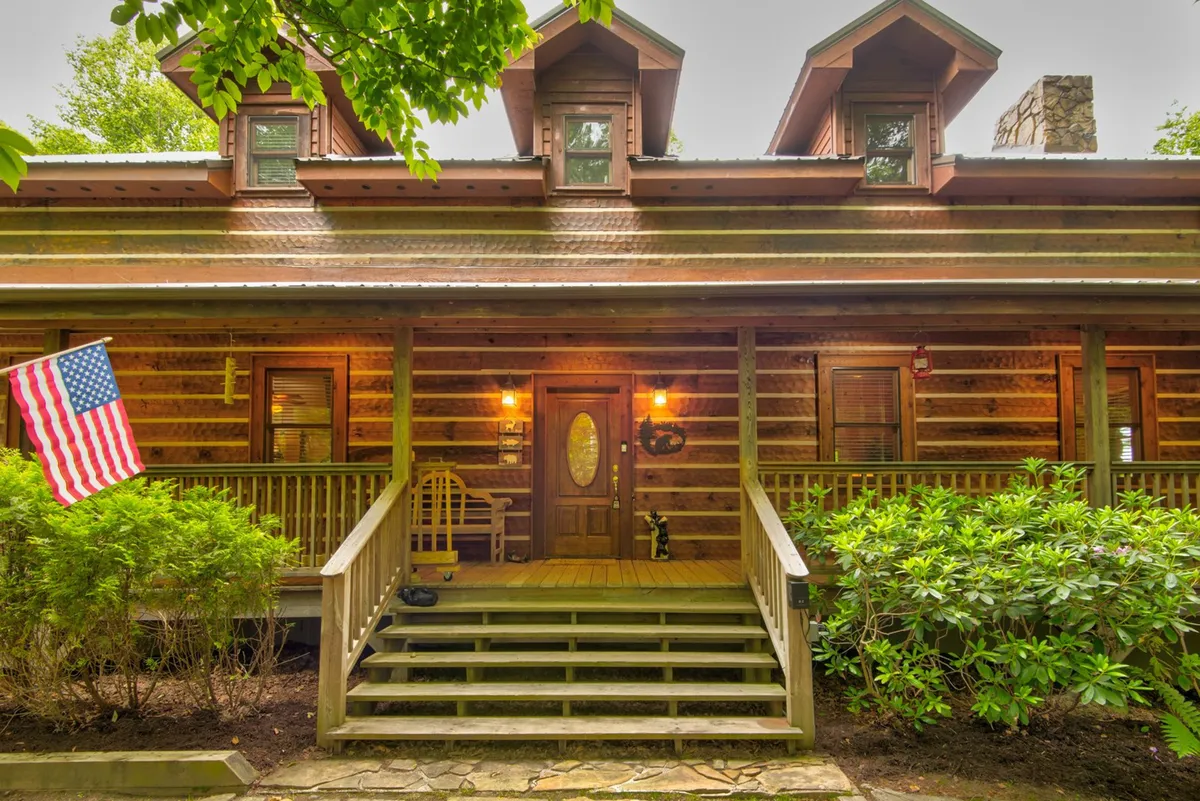 The front of the log cabin is shown with a wooden porch, American flag, and detailed log construction. Porch seating invites guests to gather before setting out to explore the local area.