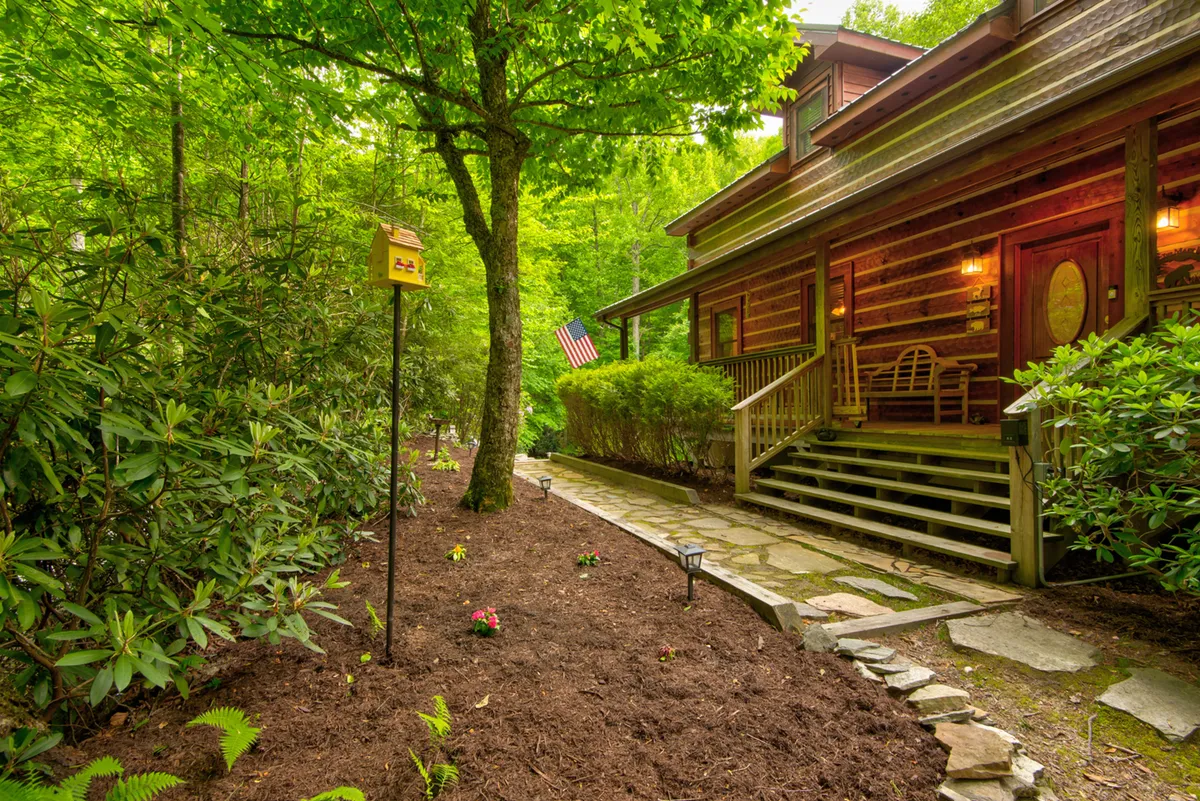 This image features the log cabin structure with a welcoming front porch, American flag, garden area, and a stone pathway leading up to the entrance. The lush greenery surrounding the home hints at the natural setting and the adventure waiting just outside.