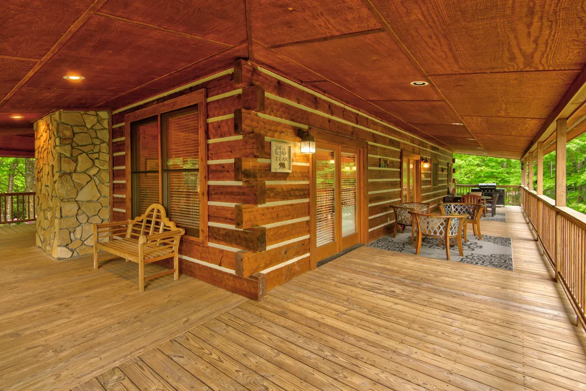 A cozy deck area with a wooden dining table and bench seating, nestled among log cabin architecture and surrounded by trees. The space is perfect for outdoor dining, enjoying fresh air, or gathering for barbecues under the stars.