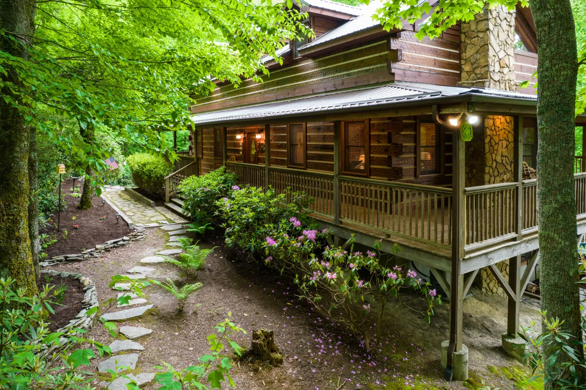A log cabin with a spacious wraparound porch nestled among lush greenery and flowering plants. The porch features comfortable seating areas, inviting guests to relax and take in the serene woodland setting.