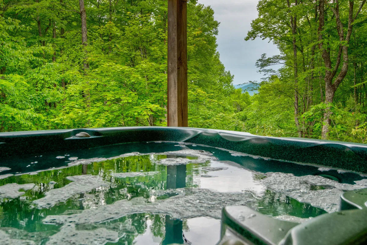This serene exterior shot centers on a bubbling outdoor hot tub surrounded by trees, with a backdrop of mountain views. The hot tub is perfectly positioned for sunset soaks or stargazing, evoking the relaxation and connection with nature described in the section.