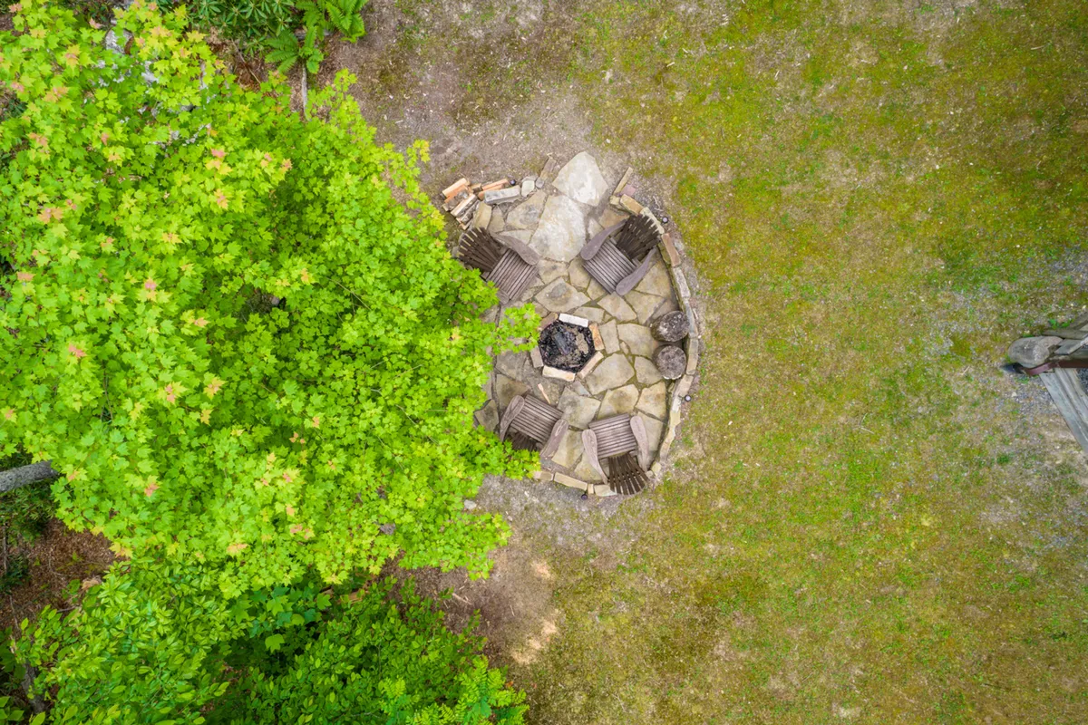 This exterior image features a cozy fire pit surrounded by comfortable seating and lush greenery. The inviting arrangement suggests a perfect spot for gathering with friends and family, sharing stories, and enjoying the warmth of the fire as evening falls. The natural surroundings emphasize the retreat’s woodland setting and sense of privacy.