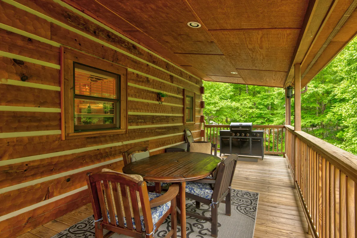 An inviting outdoor dining space with a wooden table and grill set on a deck, surrounded by greenery and a decorative rug. The area is perfect for dining, barbecuing, and gathering with family or friends.