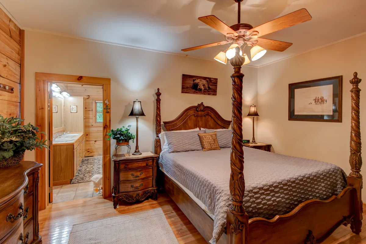 A spacious bedroom with a four-poster bed, nightstands on each side, soft lighting, and a doorway leading to an en-suite bathroom. The room features wooden accents and decorative wall art, creating a warm, restful vibe.