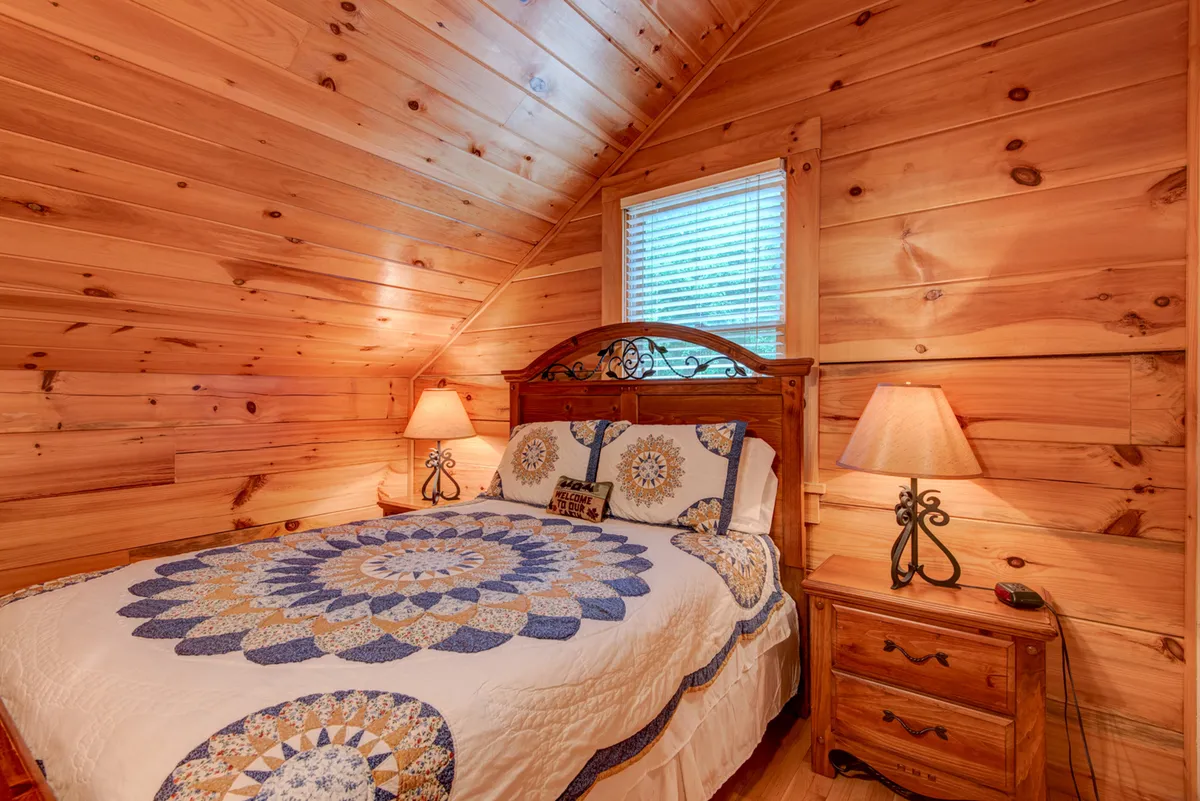 A warm, inviting bedroom with a bed dressed in a cozy quilt, wooden furniture, bedside lamps, and a window with blinds. The room exudes comfort and is perfect for quiet moments of reading or unwinding.
